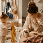 Erschöpfte Mutter sitzt morgens mit einer Tasse Kaffee am Tisch, während ein Kleinkind Kleidung hält. Warme, ruhige Alltagsszene aus dem Familienleben mit sanften Naturtönen und gemütlicher Atmosphäre.