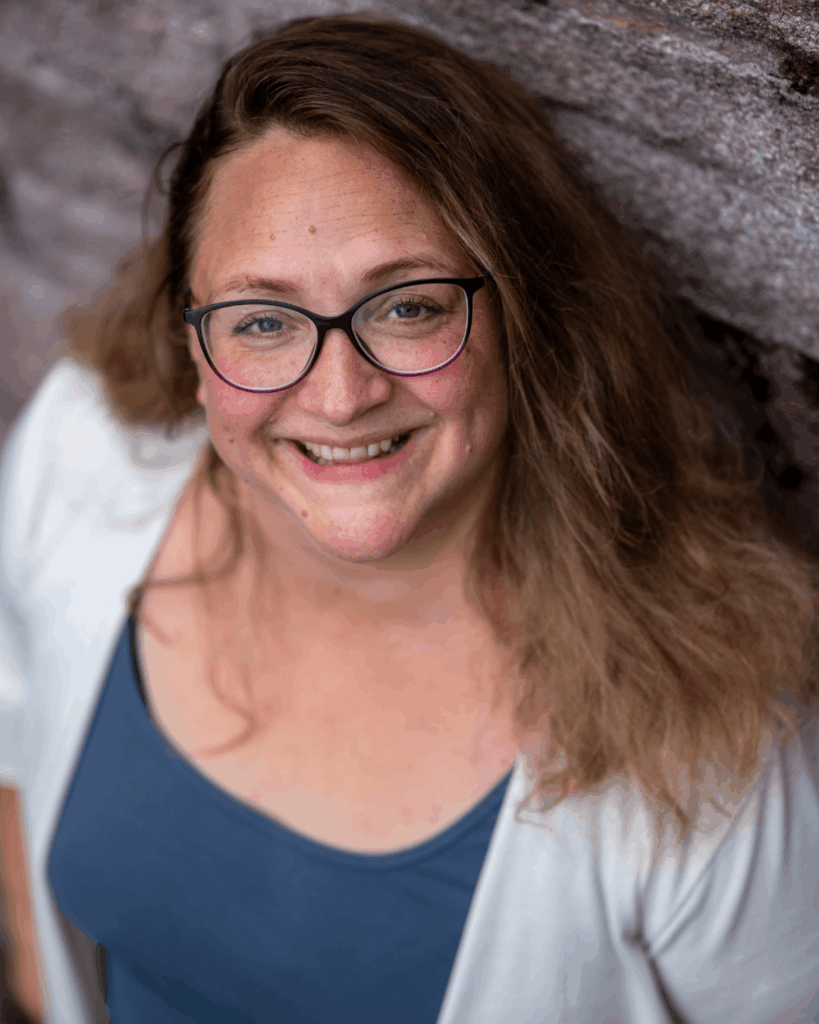 Portraitbild für Kleine Wege – Natürlich & Nahbar Portrait einer freundlich lächelnden Frau mit Brille und langen lockigen Haaren vor natürlichem Hintergrund. Warme, ruhige Bildstimmung mit natürlichem Licht und weichen Farben.