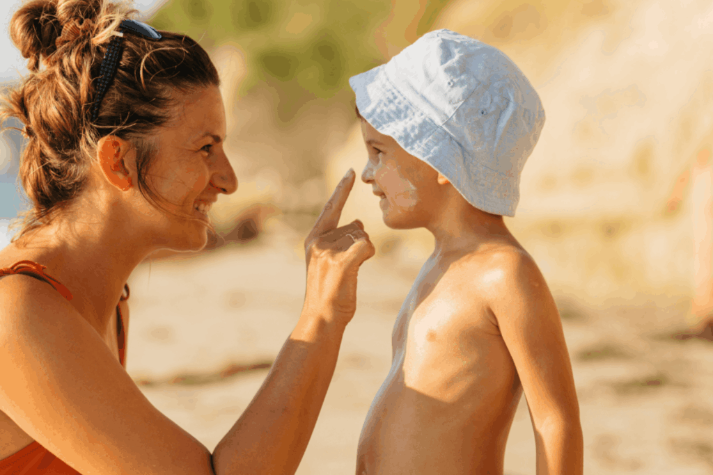 Mutter trägt ihrem Kind Sonnencreme auf Nase und Wangen auf, Kind trägt einen Sonnenhut, Strand im Hintergrund.