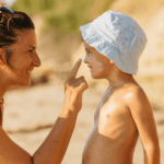 Mutter trägt ihrem Kind Sonnencreme auf Nase und Wangen auf, Kind trägt einen Sonnenhut, Strand im Hintergrund.