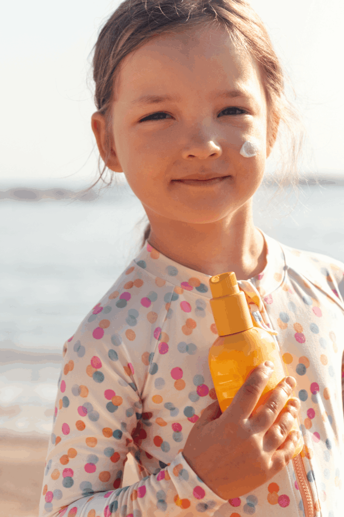 Mädchen in UV-Schutzkleidung mit gepunktetem Muster hält eine Sonnencreme-Flasche, Creme auf der Wange sichtbar, Strand im Hintergrund.