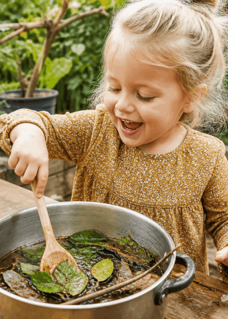 Kleinkind rührt draußen im Garten einen Topf mit Blättern, Wasser und Stöcken um