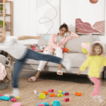 Erschöpfte Mutter mit Kopf in der Hand am Tisch, im Hintergrund tobende Kinder – Ferienstress im Familienalltag