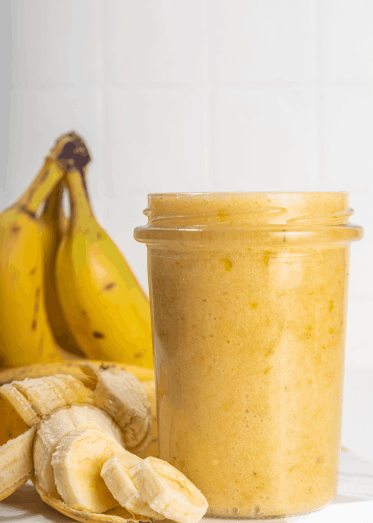 Glas mit cremiger gelber Bananenmarmelade, reife Bananen und aufgeschnittene Bananenscheiben daneben.