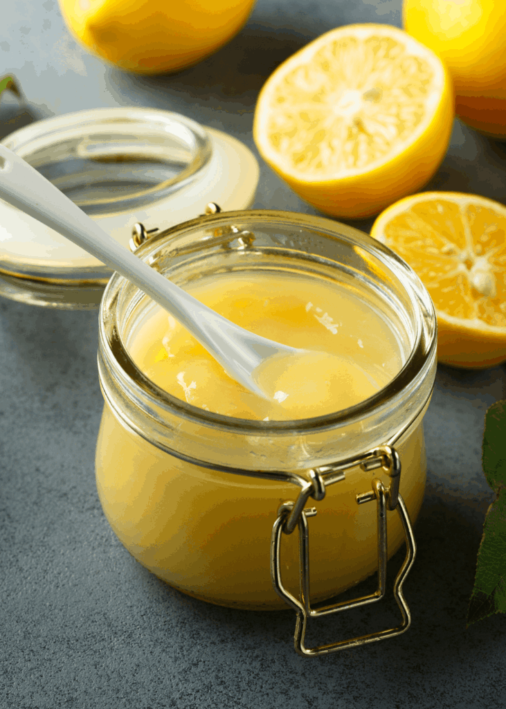 Glas mit selbstgemachtem cremigem Zitronengelee und weißem Löffel, frische Zitronenhälften im Hintergrund.