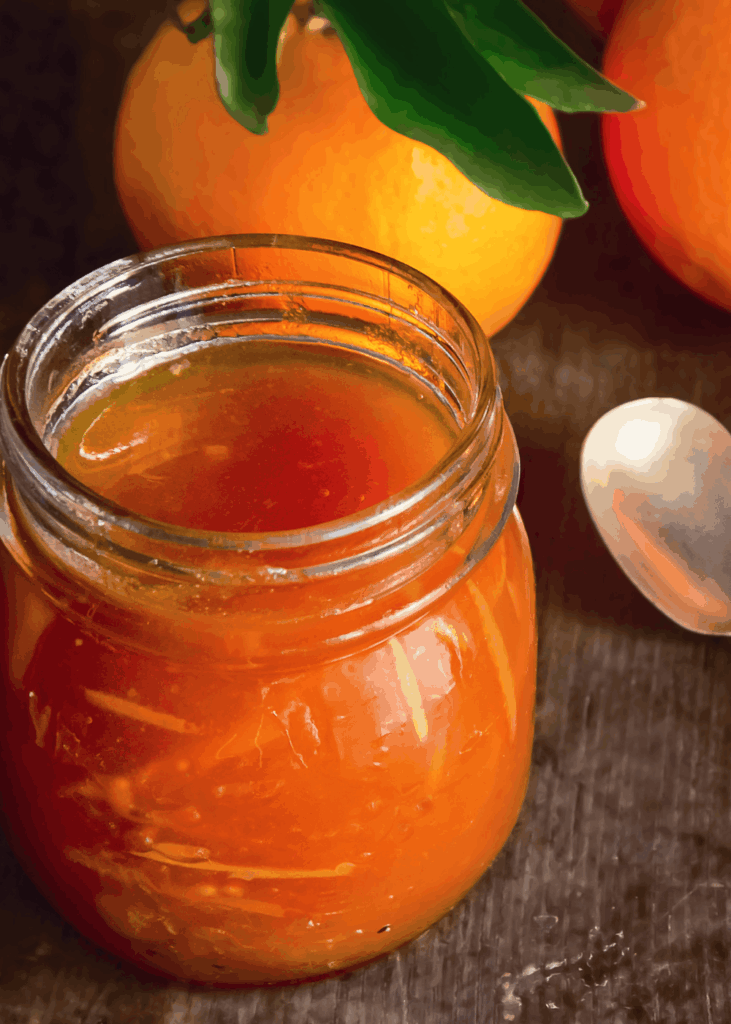 Glas mit dunkler Orangenmarmelade und Löffel daneben, frische Orangen im Hintergrund.