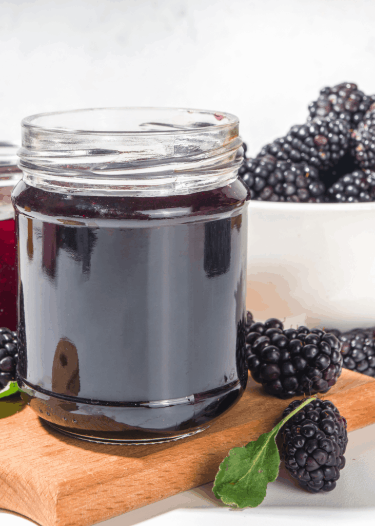 Glas mit dunkler Brombeermarmelade, frische Brombeeren und Blatt daneben.